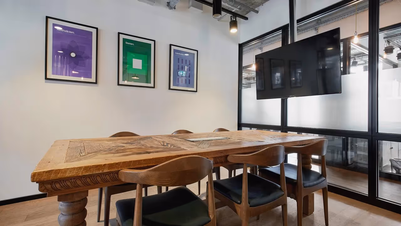 Holztisch mit sechs Stühlen, an der Wand drei bunte Bilder, Glastrennwand mit TV, Deckenbeleuchtung sichtbar.