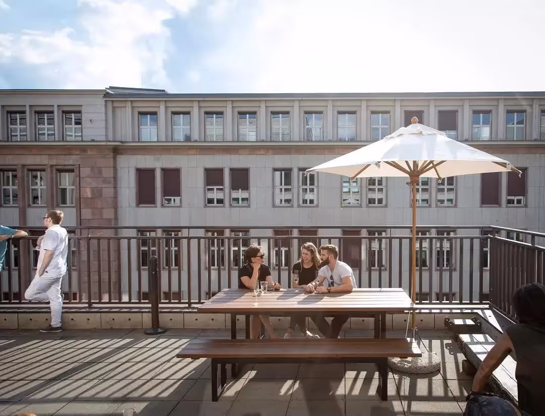 Terrasse mit Holztisch, Sonnenschirm, drei Personen sitzend, eine Person stehend, Gebäude im Hintergrund.