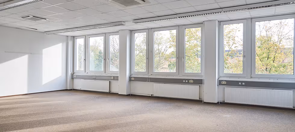 Leerer Raum mit Teppichboden, großen Fenstern, weißen Wänden, Deckenplatten und Heizkörpern unter den Fenstern.