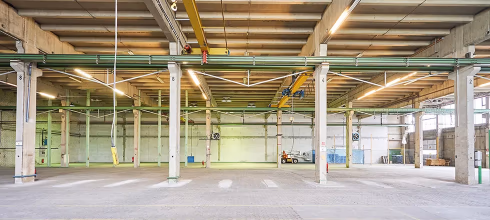 Weitläufige Industriehalle mit Betonpfeilern, Deckenbeleuchtung und gelbem Kran, leere offene Fläche.