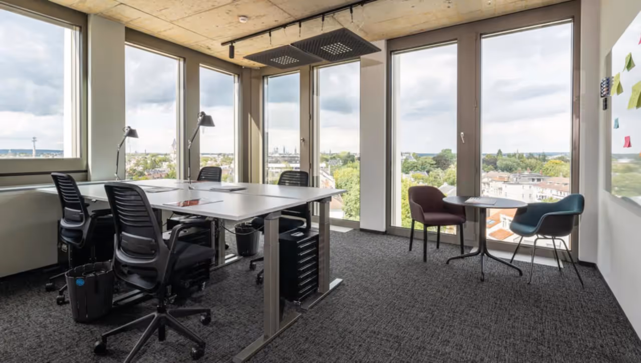 Büro mit weißen Schreibtischen, schwarzen Stühlen, grauem Teppich, Panoramafenstern und kleinem Sitzplatz.