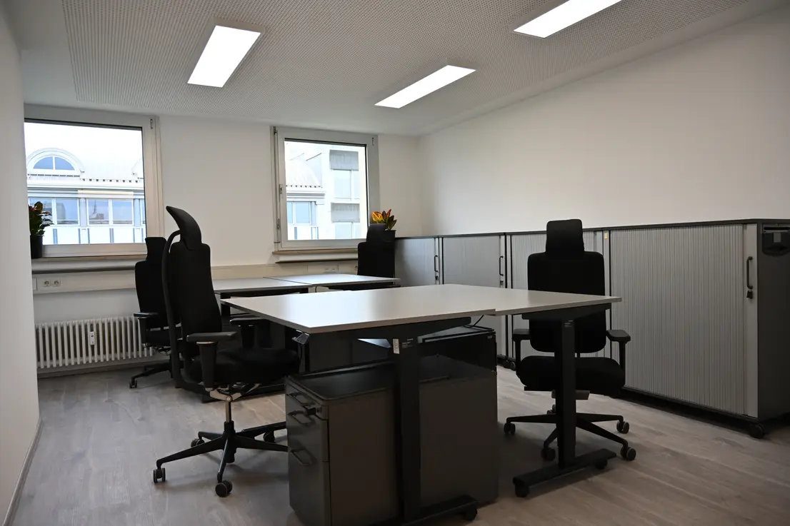 Heller Büroraum mit vier schwarzen Bürostühlen, weißen Tischen, großen Fenstern und grauen Rollcontainern.