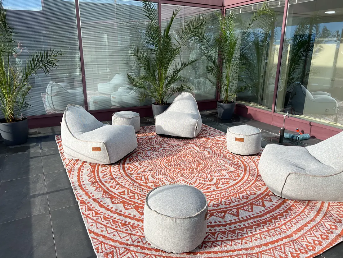 Graue Sitzsäcke und Hocker auf rotem Musterteppich, umgeben von Palmen in Töpfen auf einer Terrasse.