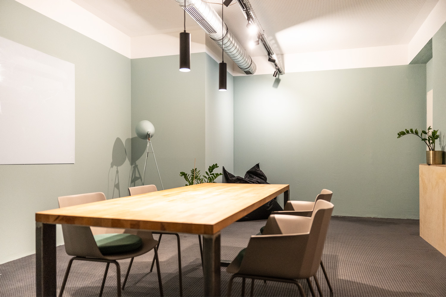 Großer Holztisch mit sechs Stühlen, grüne Wände, Deckenleuchten, Pflanzen, Stehlampe, Teppichboden, Lüftungsrohr.