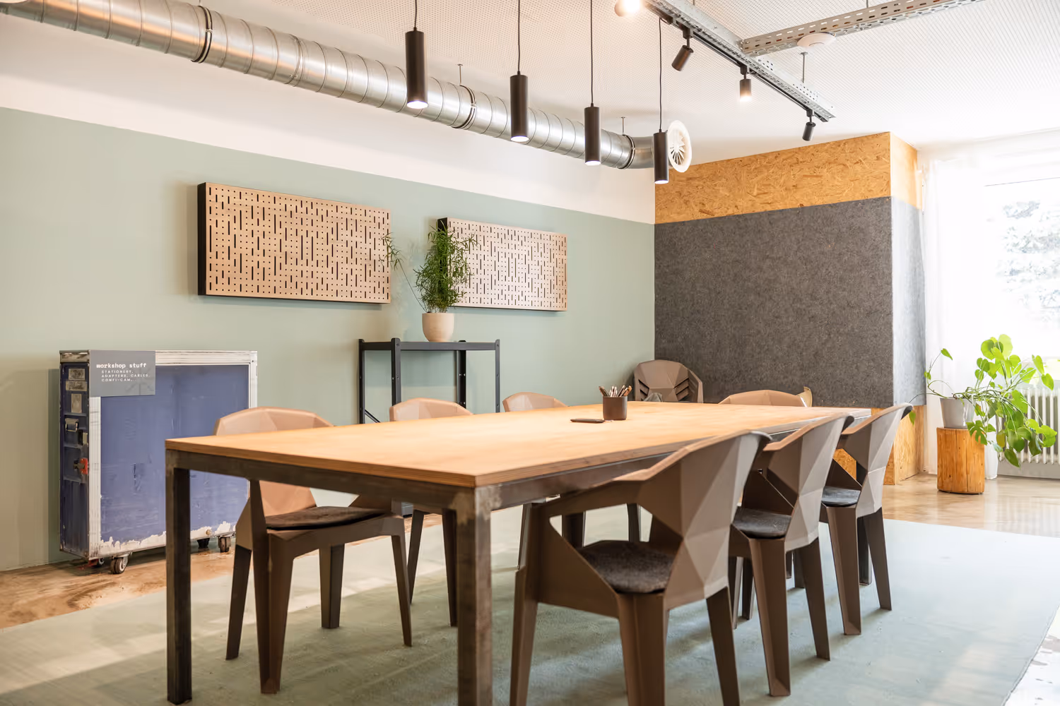 Holztisch mit geometrischen Stühlen, hängende Lampen, grüne Wand mit Pflanzen, Holz- und Filzverkleidung.