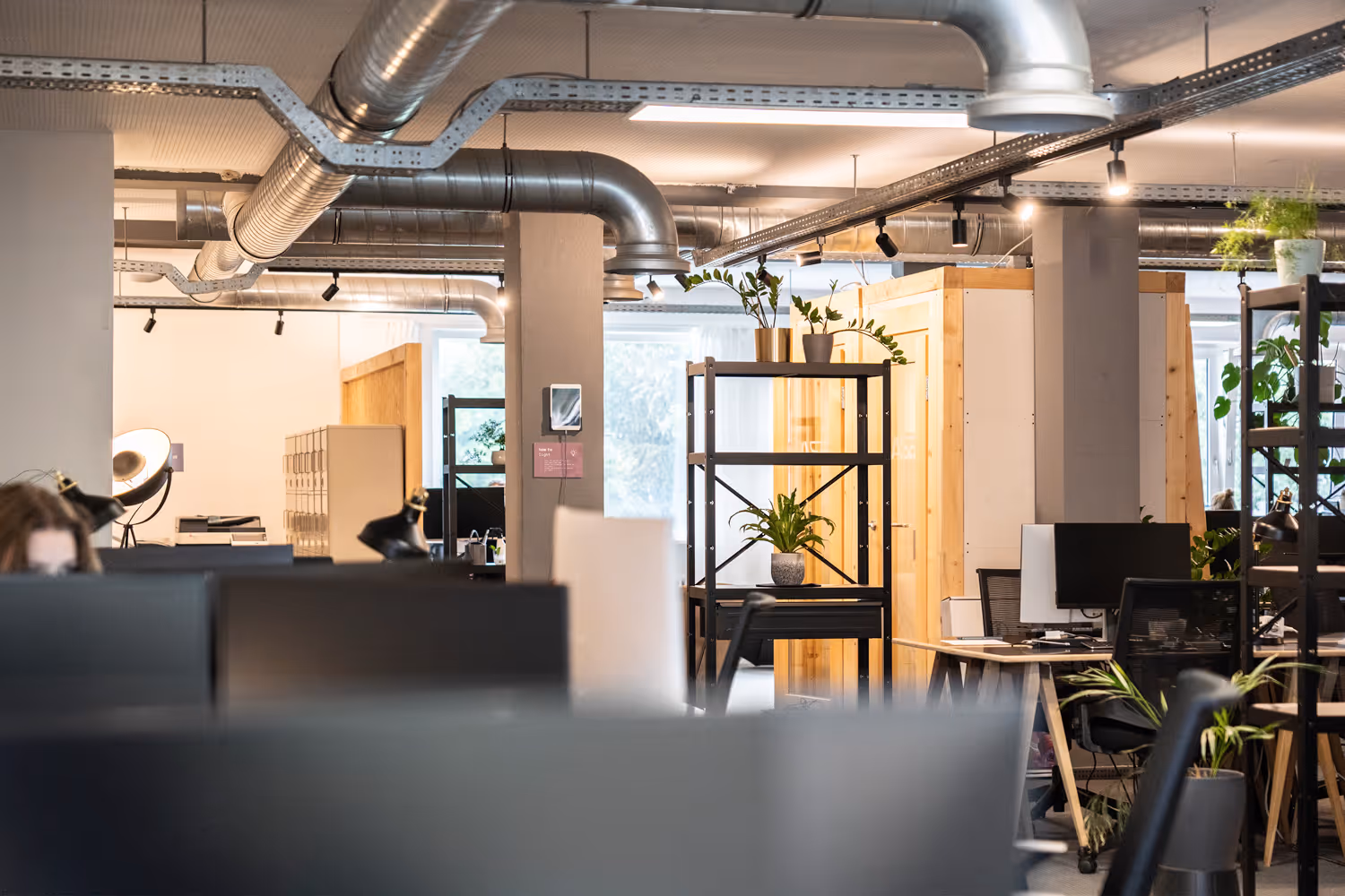 Industrieller Coworking-Raum mit Pflanzen, Holztrennwänden, Schreibtischen, Lampen und sichtbaren Lüftungsrohren.