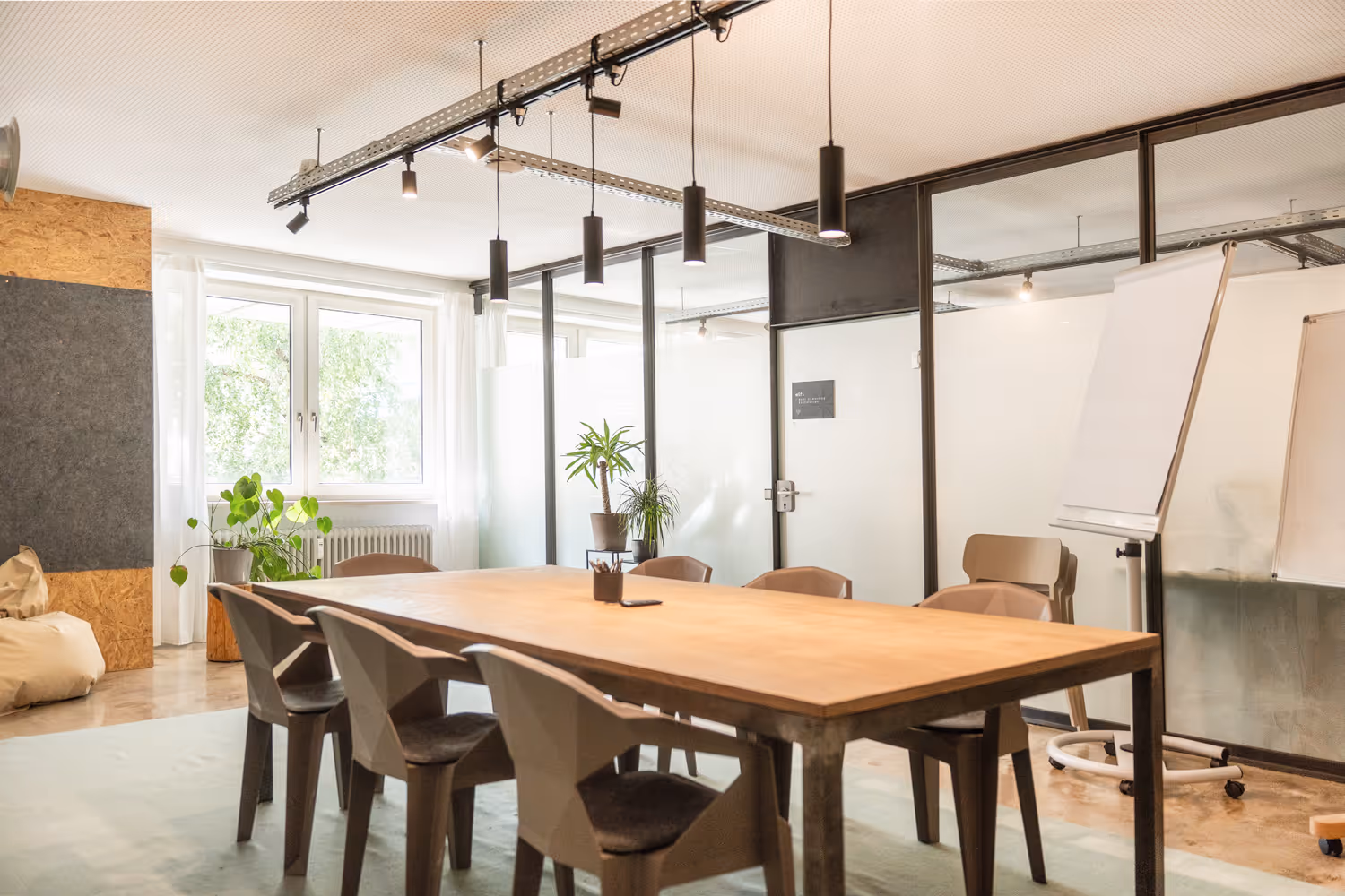 Heller Besprechungsraum mit Holztisch, modernen Stühlen, Pflanzen, großen Fenstern und Schiebetür aus Glas.