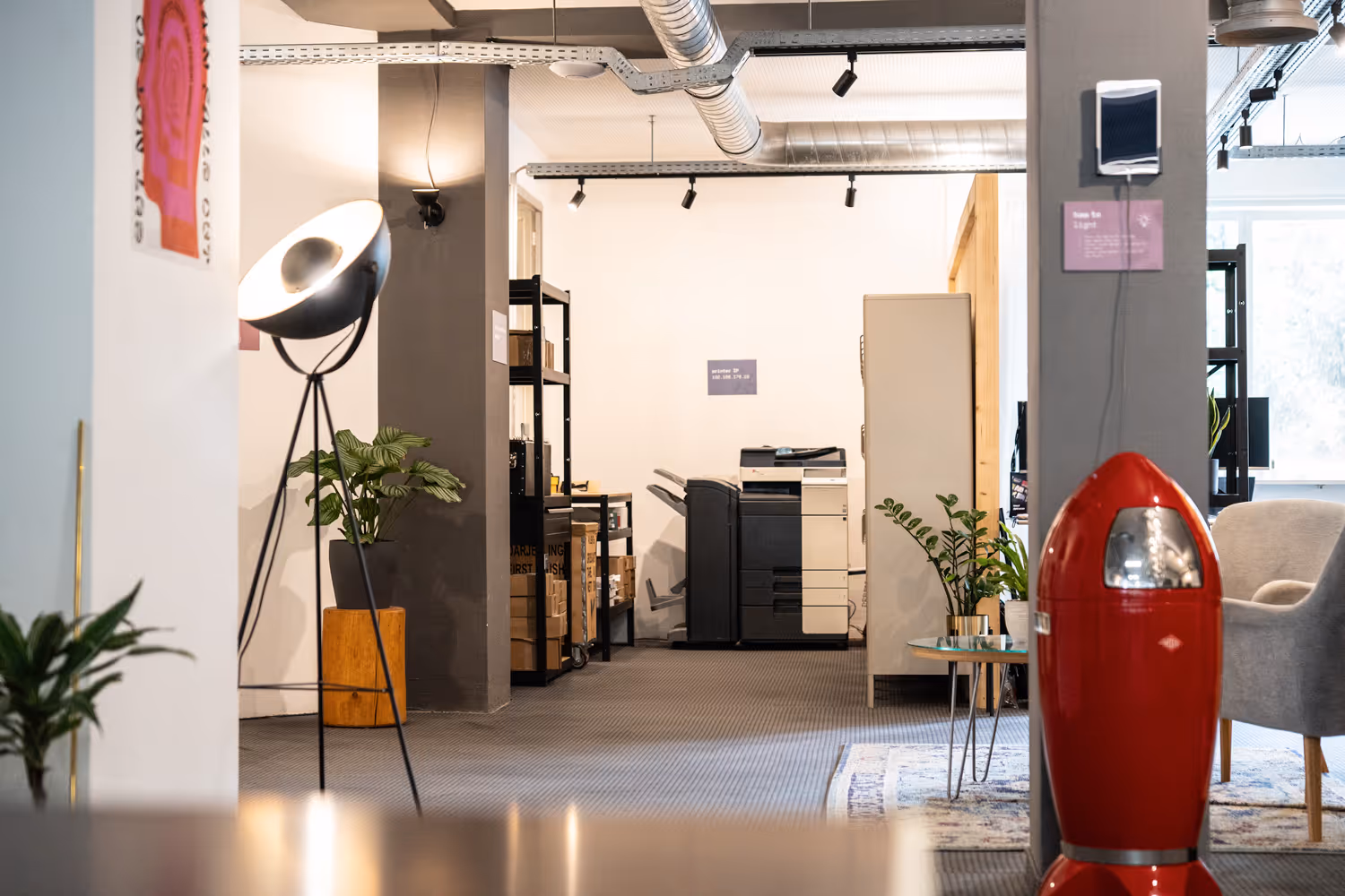 Coworking-Bereich mit Stehlampe, Drucker, Pflanzen, grauem Sessel, rotem Mülleimer, Regalen, Teppich und Lüftungsrohren.