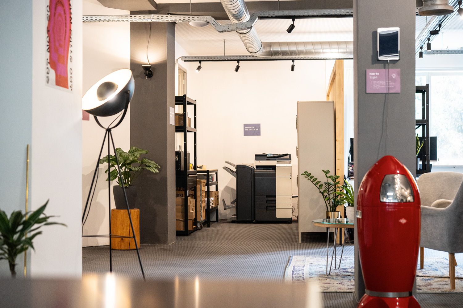 Coworking-Bereich mit Stehlampe, Drucker, Pflanzen, grauem Sessel, rotem Mülleimer, Regalen, Teppich und Lüftungsrohren.