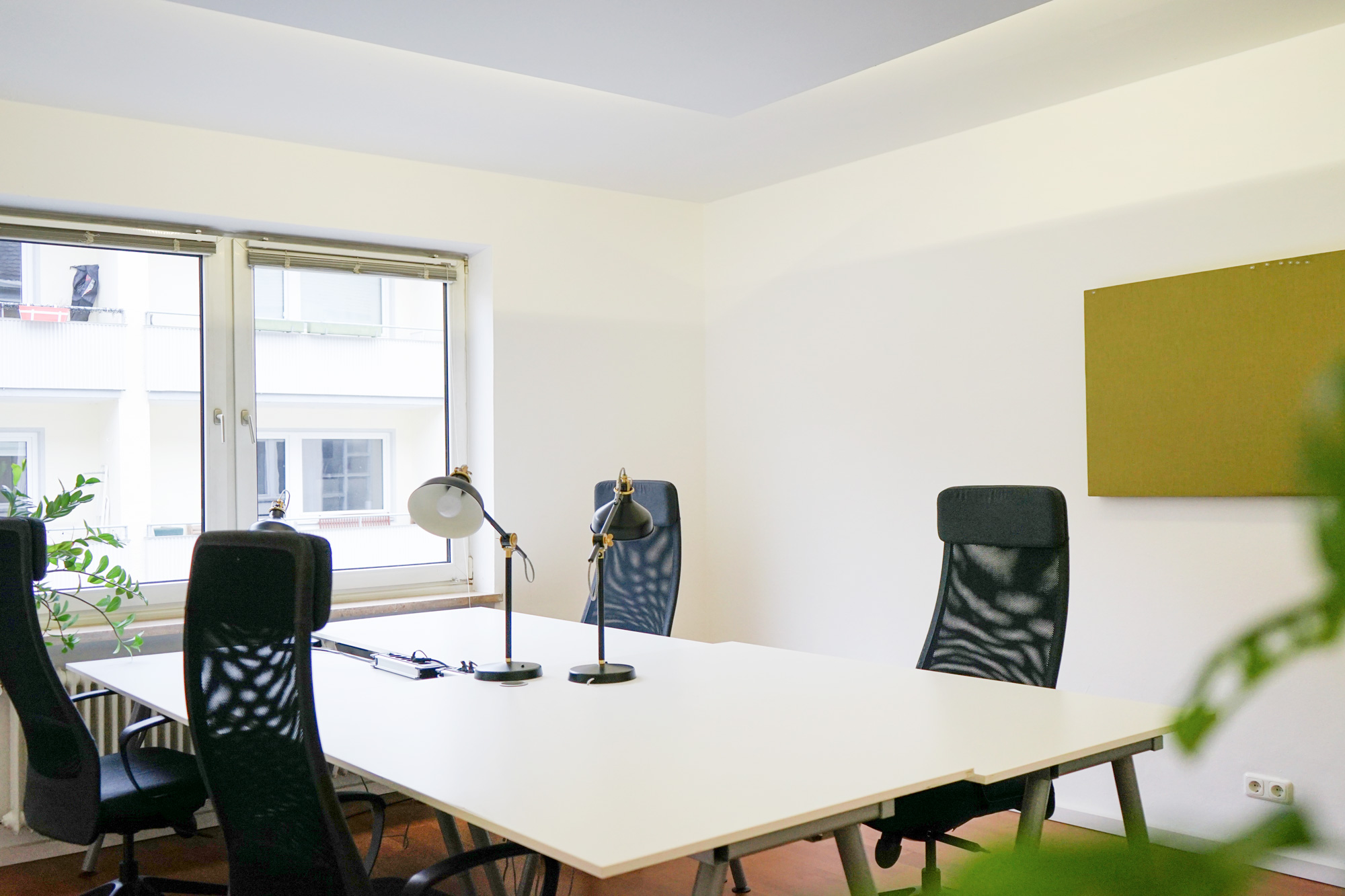 Heller Raum mit großem weißen Tisch, vier schwarzen Bürostühlen, zwei Lampen und Fenster mit Blick nach draußen.