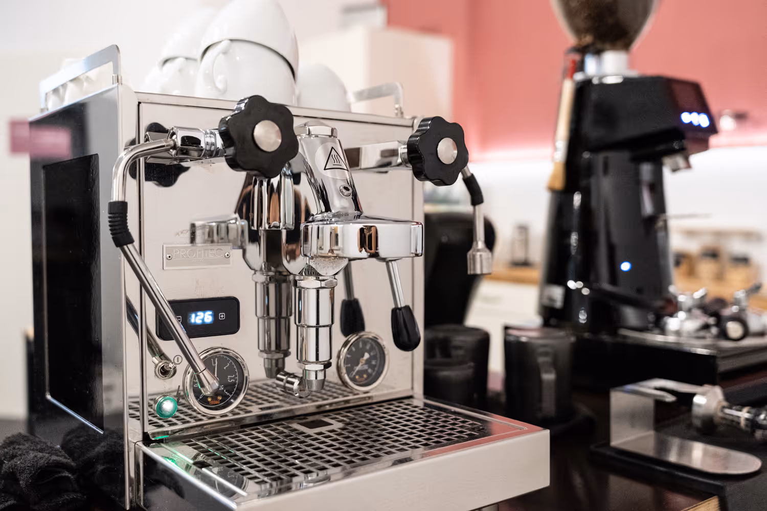 Silberne Espressomaschine mit Tassen darüber, daneben schwarze Kaffeemühle in modernem Raum mit rosa Wand.