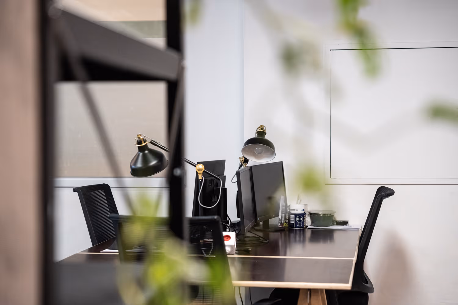 Arbeitsplatz mit zwei Monitoren, schwarzen Bürostühlen, grünen Schreibtischlampen und Pflanzen im Vordergrund.