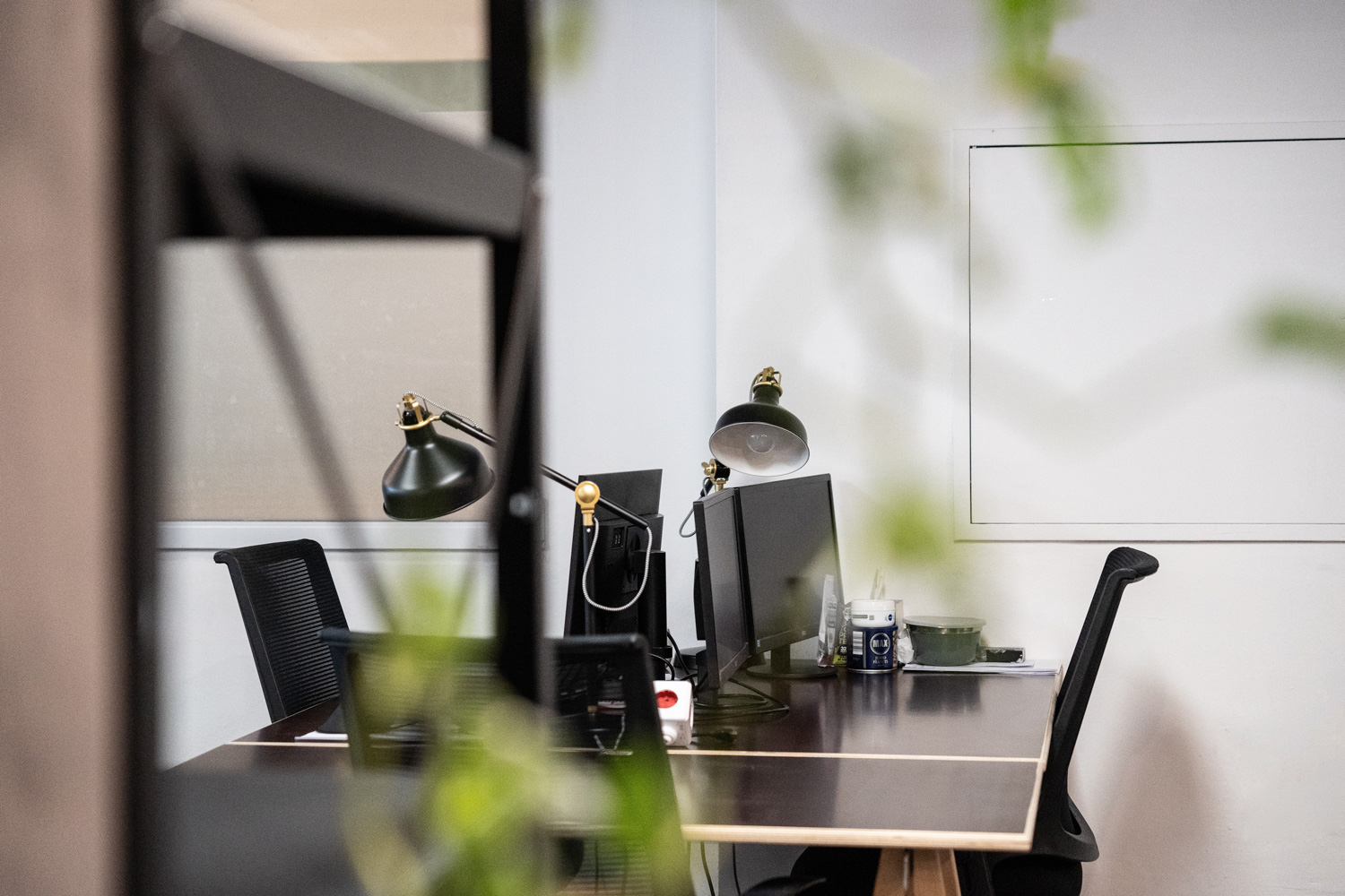 Arbeitsplatz mit zwei Monitoren, schwarzen Bürostühlen, grünen Schreibtischlampen und Pflanzen im Vordergrund.