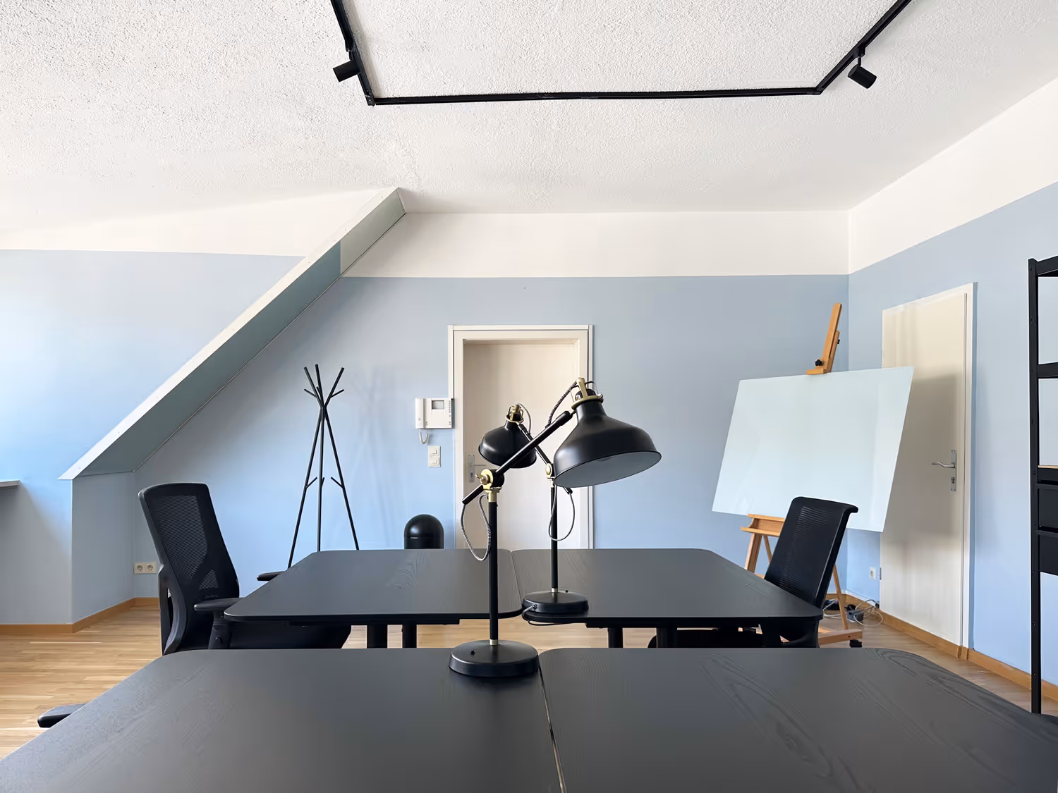 Schwarze Tische, Bürostühle, Stehlampe, Staffelei mit Glasplatte, blassblaue Wände, schräges Dachfenster, Kleiderständer.