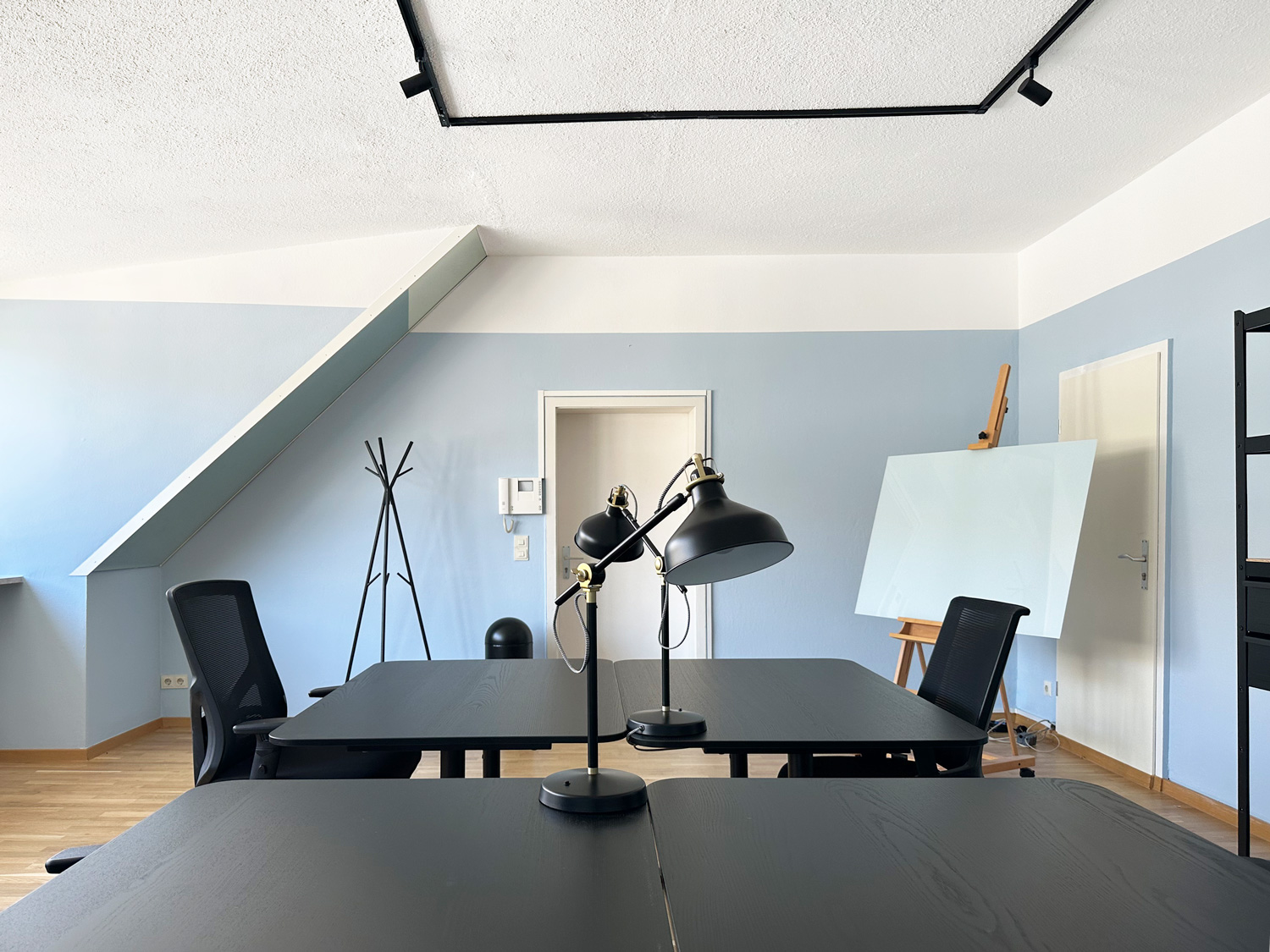 Schwarze Tische, Bürostühle, Stehlampe, Staffelei mit Glasplatte, blassblaue Wände, schräges Dachfenster, Kleiderständer.