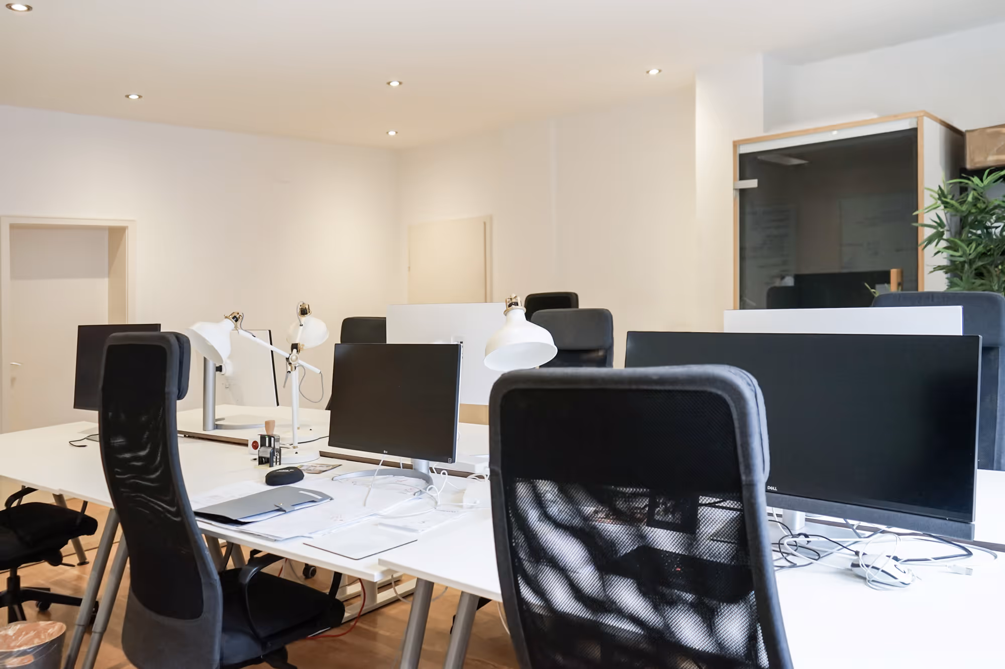 Weiße Schreibtische mit schwarzen Monitoren, ergonomische Stühle, weiße Lampen, helle Wände, Glaskabine, Pflanzen.