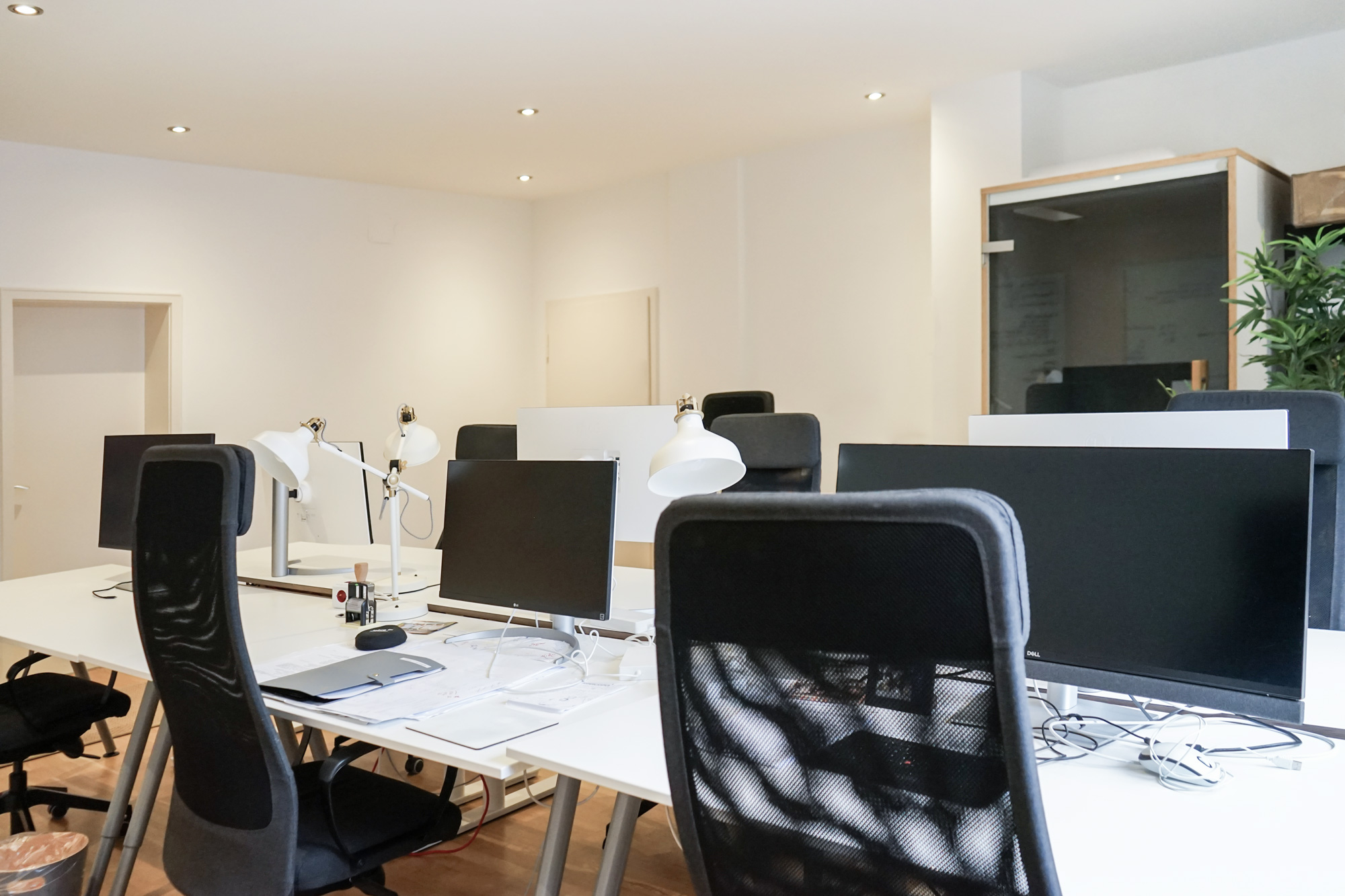 Weiße Schreibtische mit schwarzen Monitoren, ergonomische Stühle, weiße Lampen, helle Wände, Glaskabine, Pflanzen.