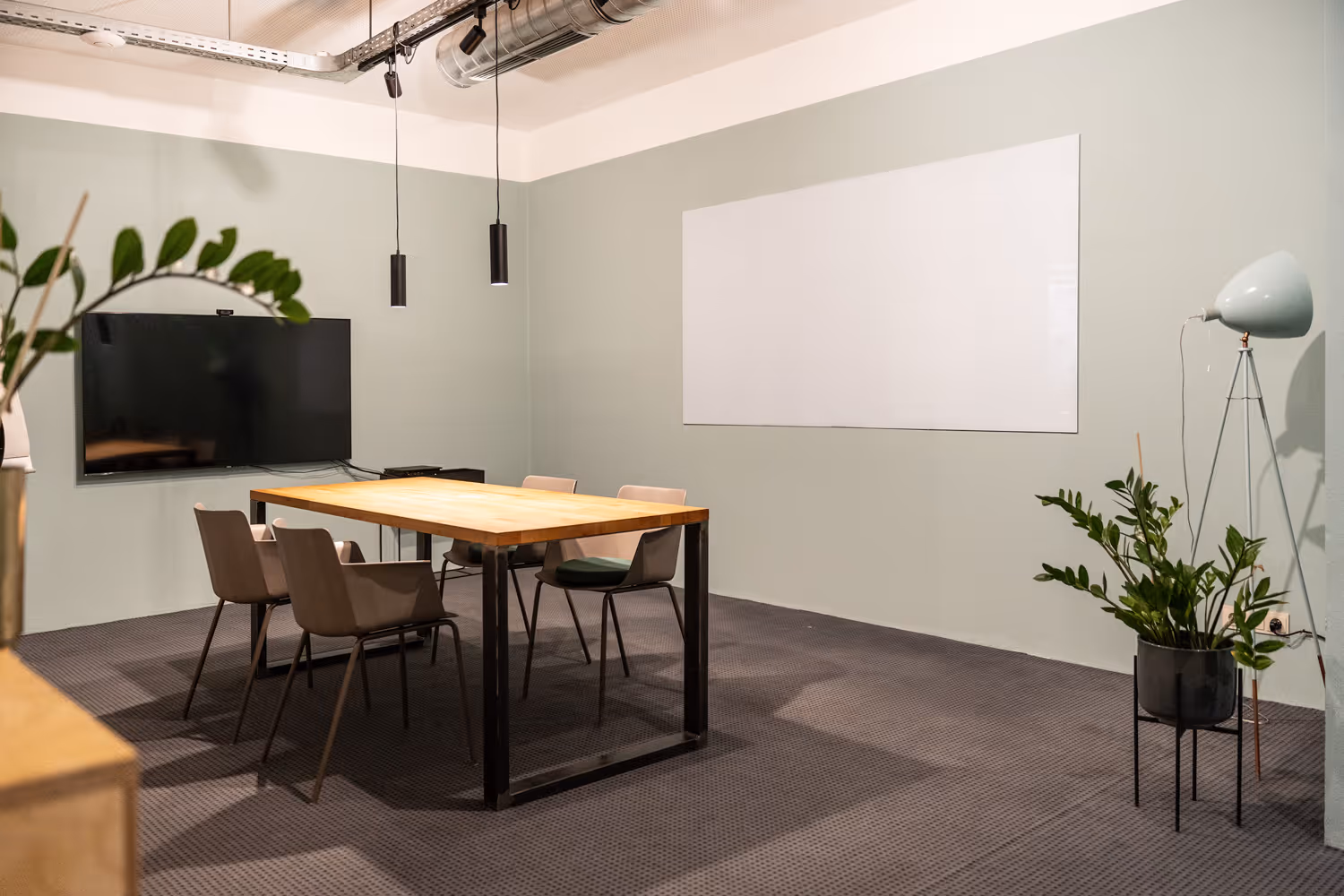 Holztisch mit Stühlen, TV an grüner Wand, weißes Board, Hängelampen, Pflanze in Ecke, grauer Teppichboden.