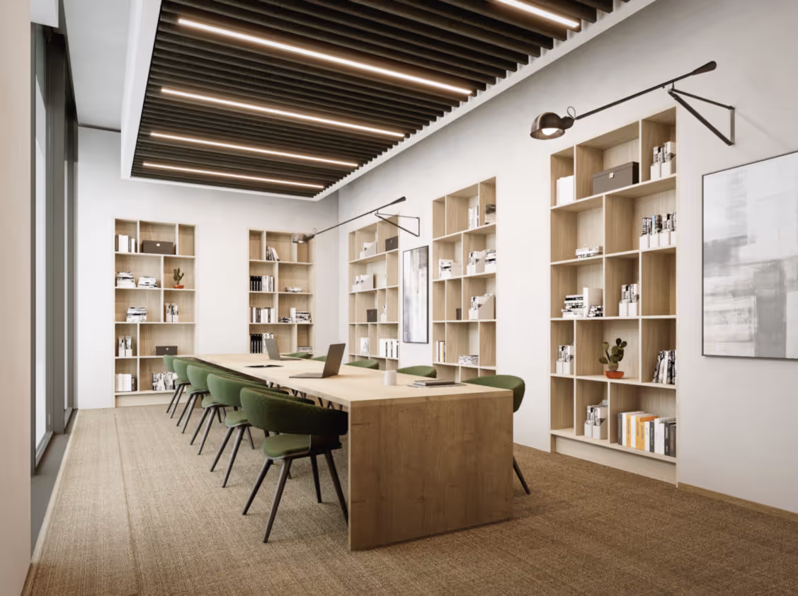 Langer Holztisch mit grünen Stühlen, Regale mit Büchern, moderne Beleuchtung, heller Raum mit Teppichboden.