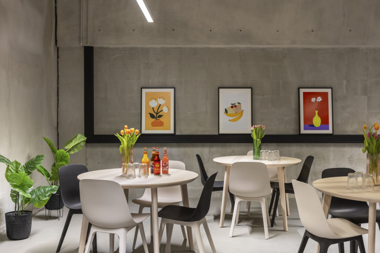Runde Tische mit weißen Stühlen, Tulpenvasen, bunte Wandbilder, Betonwände, moderne Hängelampe, Pflanzen.