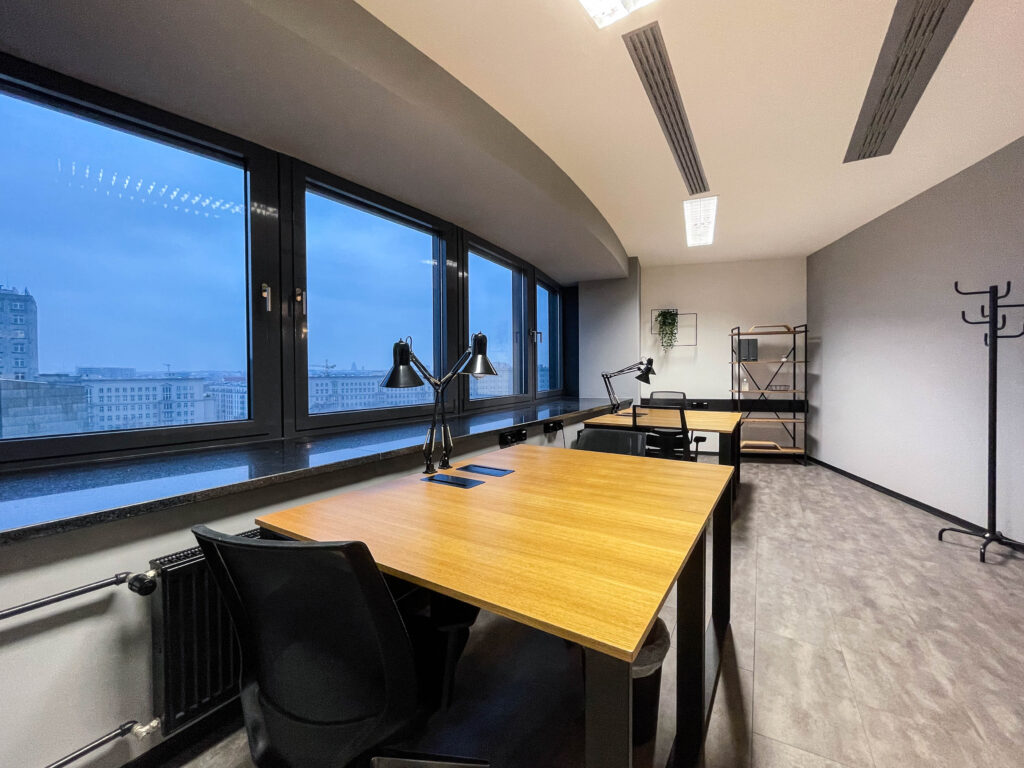 Großer Raum mit Holztischen, schwarzen Stühlen, Schreibtischlampen, Fensterfront und grauem Bodenbelag.