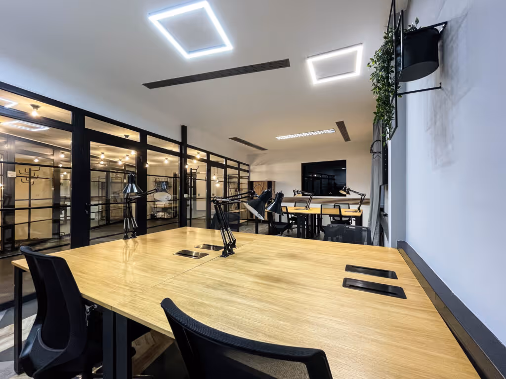 Helle Bürofläche mit Holztischen, schwarzen Stühlen, Tischlampen, Glaswänden und modernen Deckenleuchten.