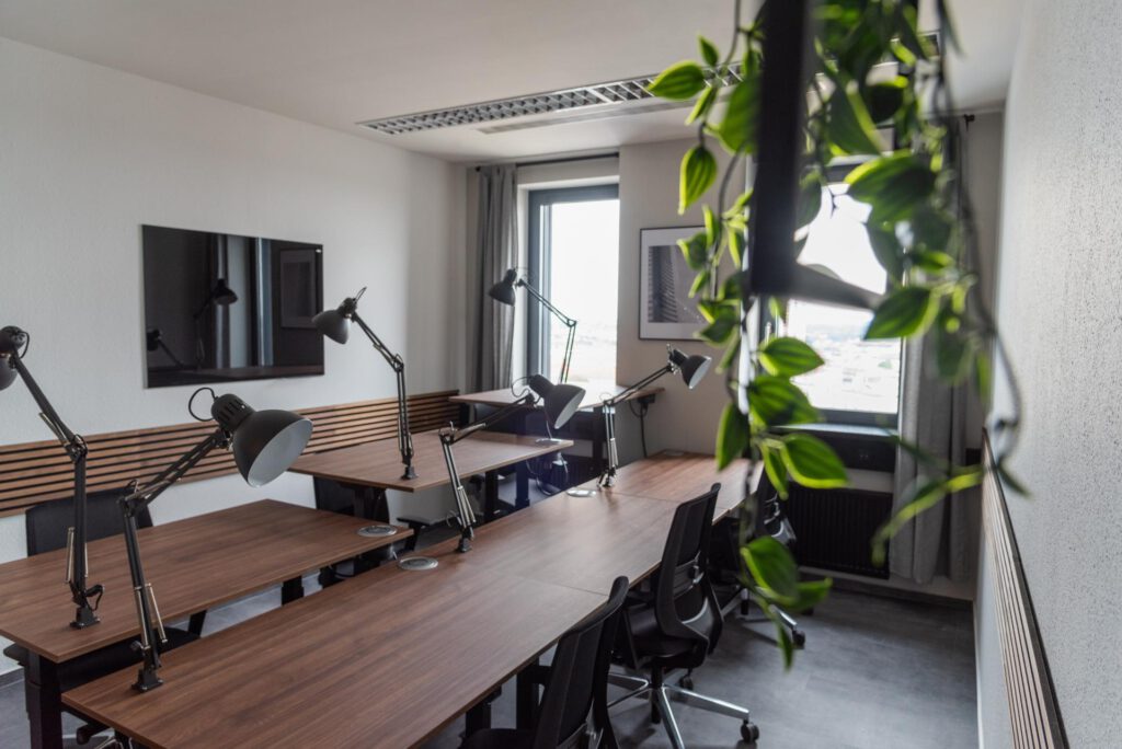 Holztische mit schwarzen Stühlen, Schreibtischlampen, Wandbild, Fenster mit Vorhängen, hängende Pflanze.