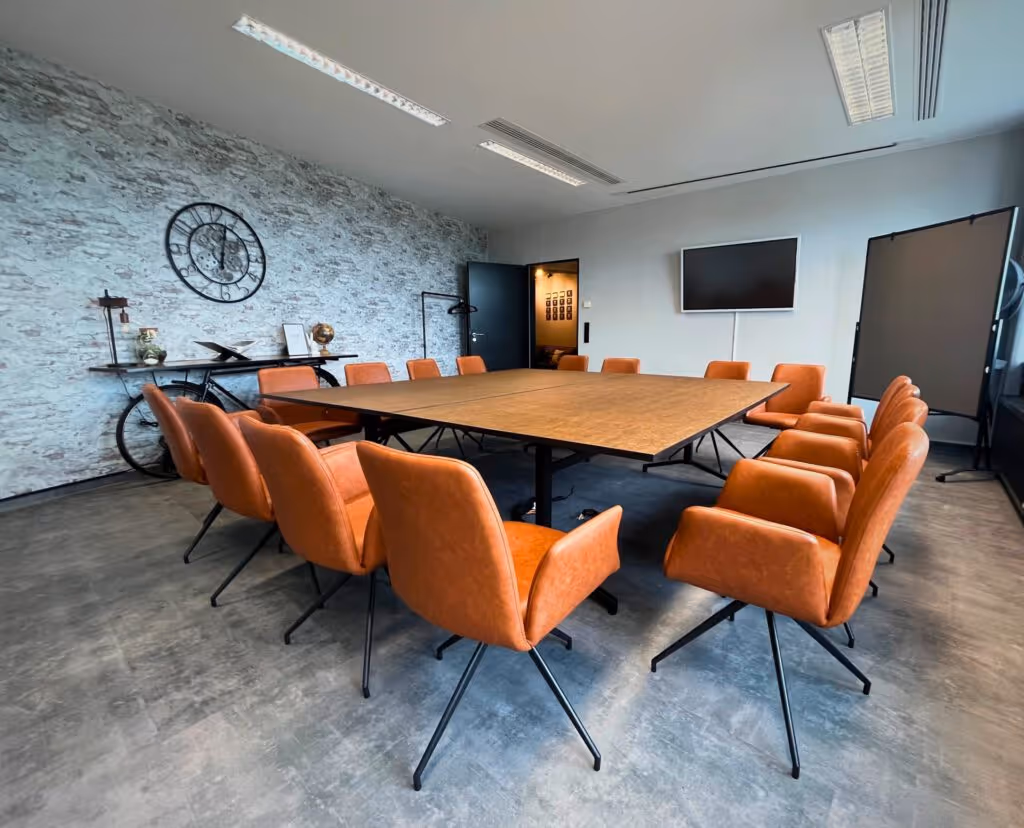 Konferenzraum mit großem Tisch, orangefarbenen Stühlen, Steinwand, Uhr, TV-Bildschirm und Lampe.