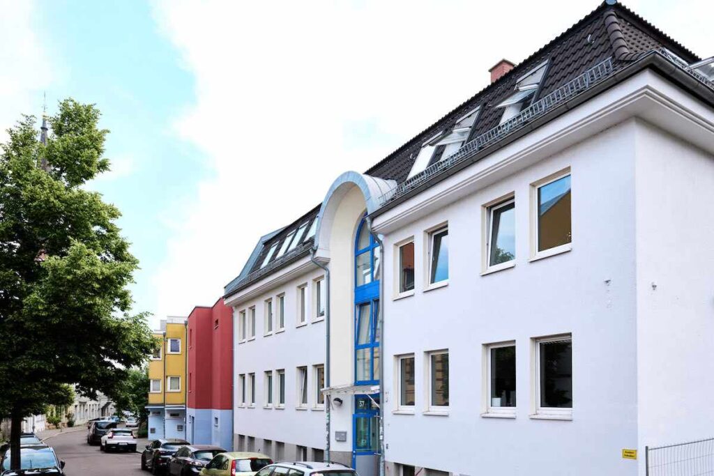 Weiße Fassade mit blauen Akzenten, Dachfenstern, Baum links, parkende Autos rechts, bunte Nachbargebäude.