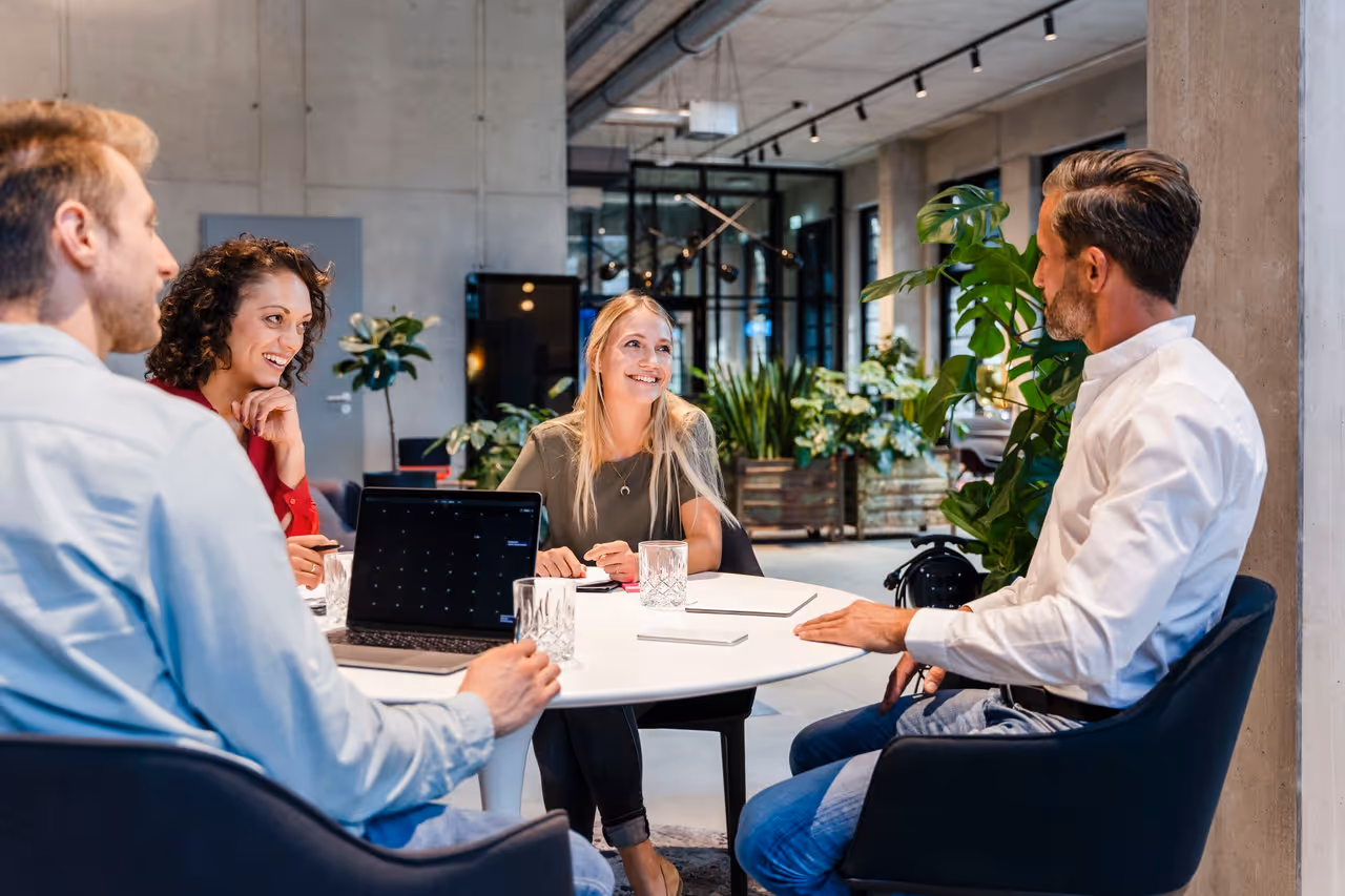 Vier Personen sitzen an einem runden Tisch in modernem Büro, umgeben von Pflanzen und Betonwänden.