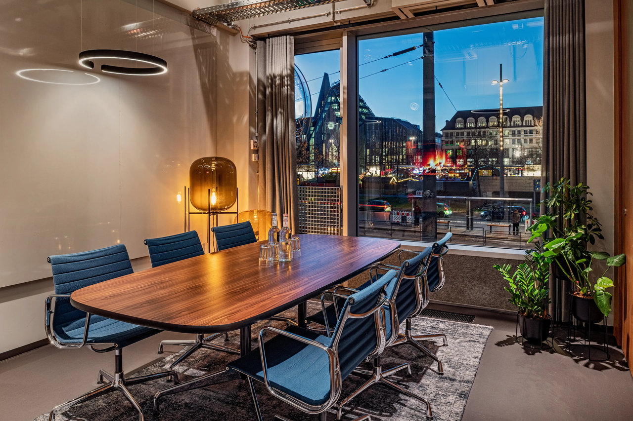 Konferenztisch mit blauen Stühlen, Holzplatte, Stehlampe, Fensterblick auf Stadt, Pflanzen, moderne Beleuchtung.