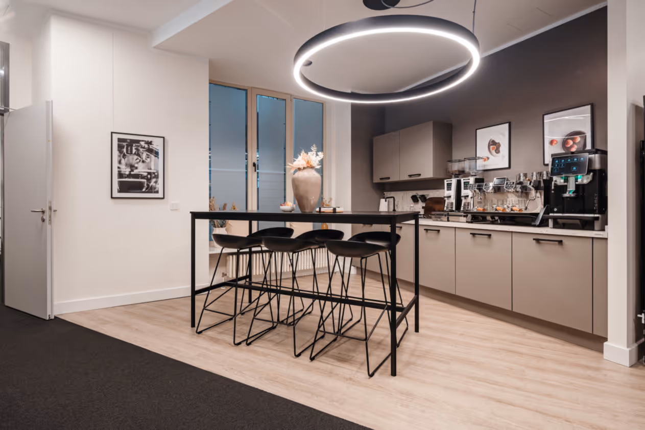 Heller Raum mit Bartisch, schwarzen Hockern, Küche, Kaffeemaschine, großem Ringlicht und Holzfußboden.