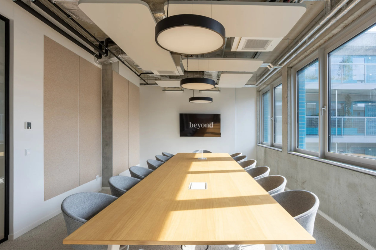 Langer Holztisch mit grauen Stühlen, große Fenster, moderne Deckenlampen, Bildschirm mit "beyond" an der Wand.