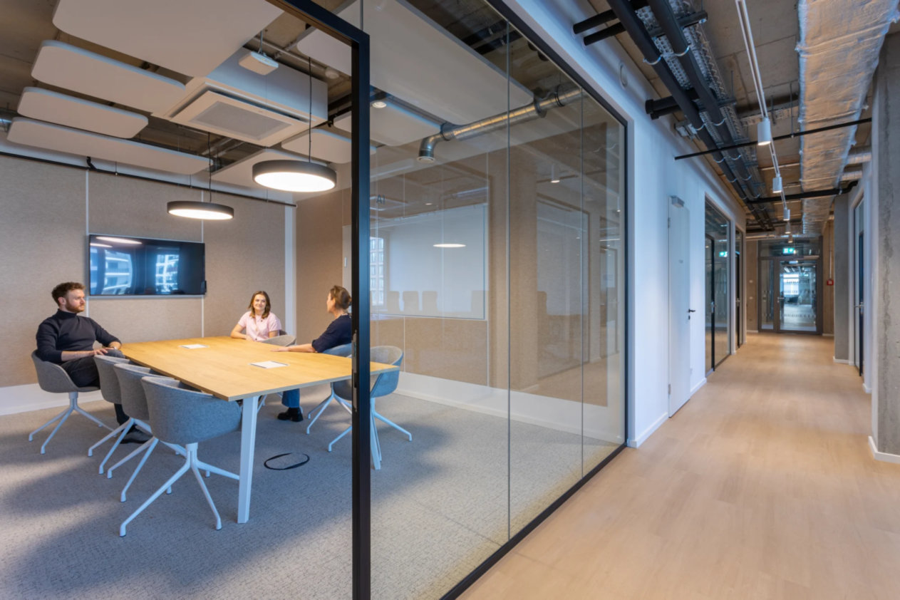 Besprechungsraum mit Holztisch, grauen Stühlen, drei Personen, Glaswänden, Deckenbeleuchtung, sichtbare Lüftung.