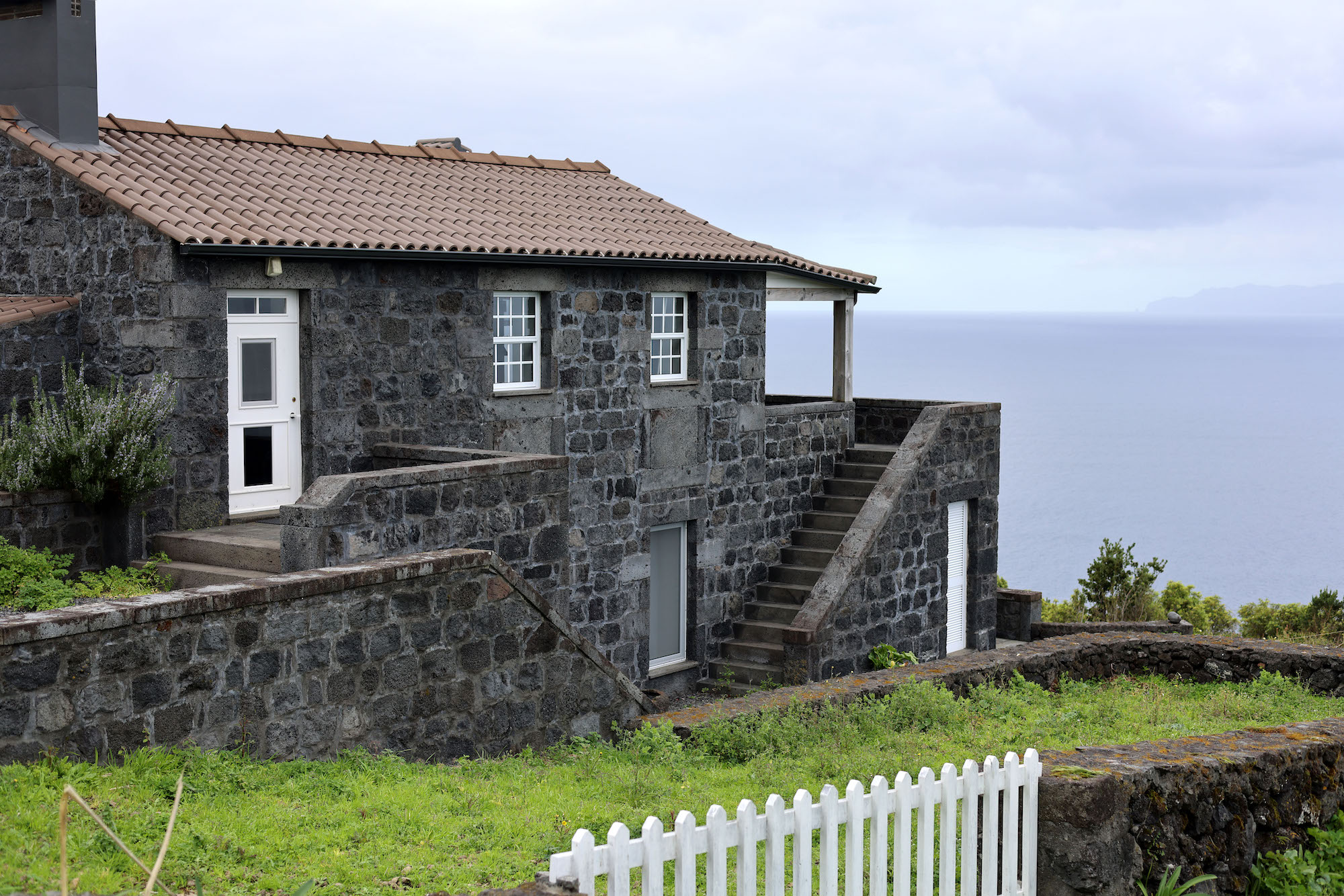 Stone house with a tiled roof, exterior stairs, and ocean view in the background.