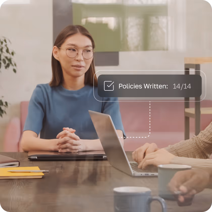 Woman with glasses sitting at a table across from someone typing on a laptop with a graphic showing 'Policies Written: 14/14'.