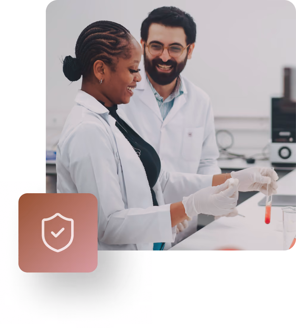 Two scientists in white lab coats and gloves examining a test tube with red liquid in a laboratory, smiling as they work together.