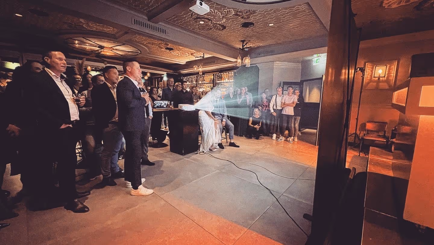 Een grote groep bruiloftgasten kijkt naar een scherm waar met een beamer iets op geprojecteerd wordt.