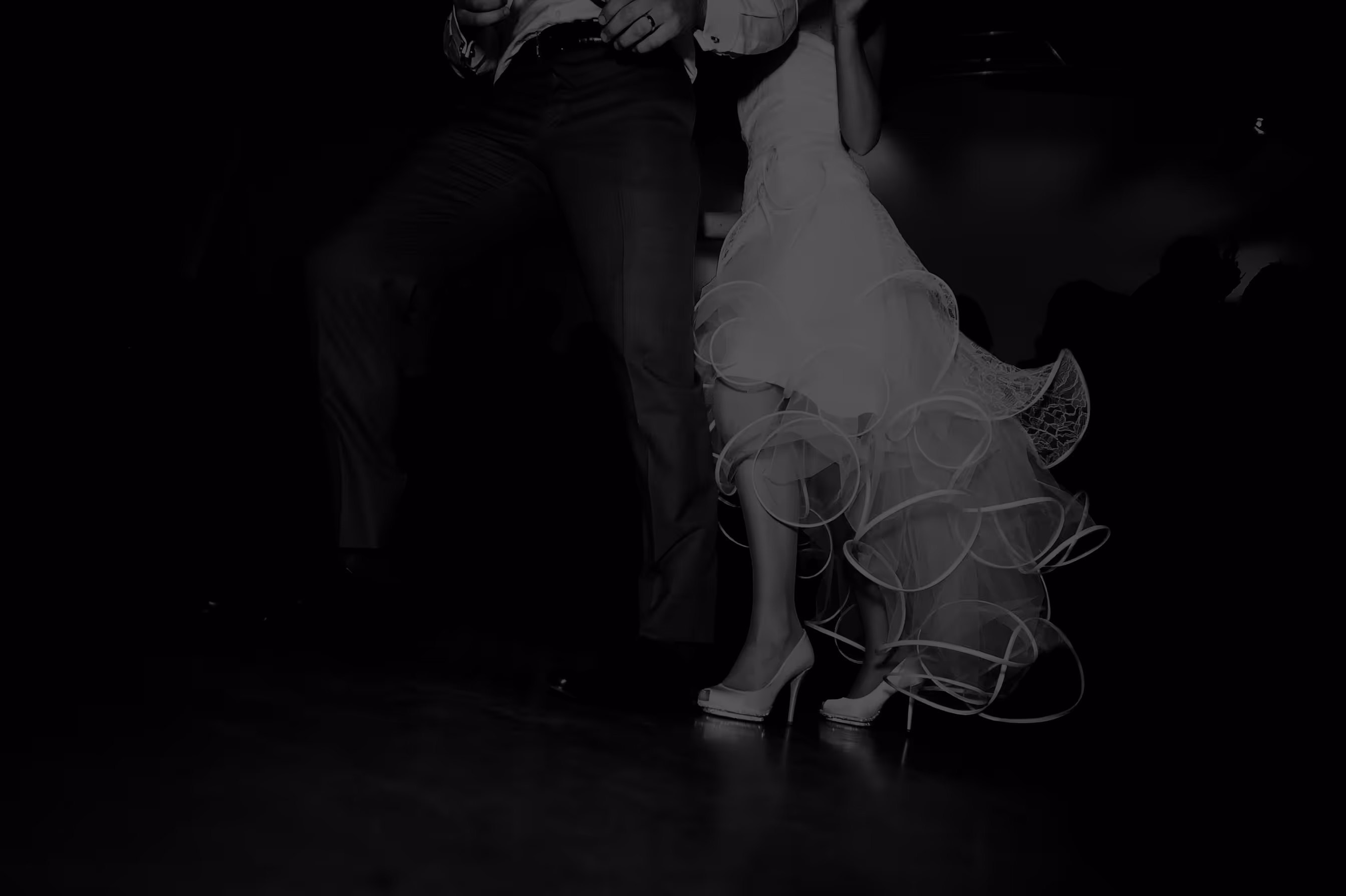 De benen van een dansend bruidspaar, waarbij de man een pak draagt en de bruid een witte jurk met laagjes en hoge witte hakken.