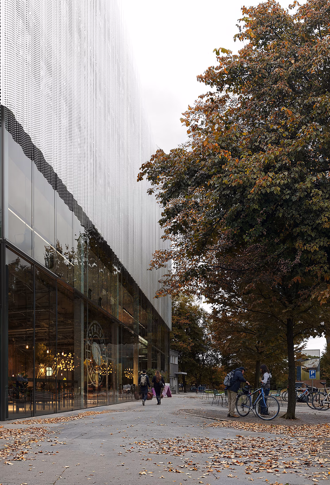 Architectural visualization of a modern logistics hub in Switzerland, featuring a perforated metal façade and a transparent ground floor activating the urban streetscape. The autumn setting, with fallen leaves and cyclists, enhances the integration of sustainable mobility and contemporary design. A high-end render showcasing innovative commercial architecture.