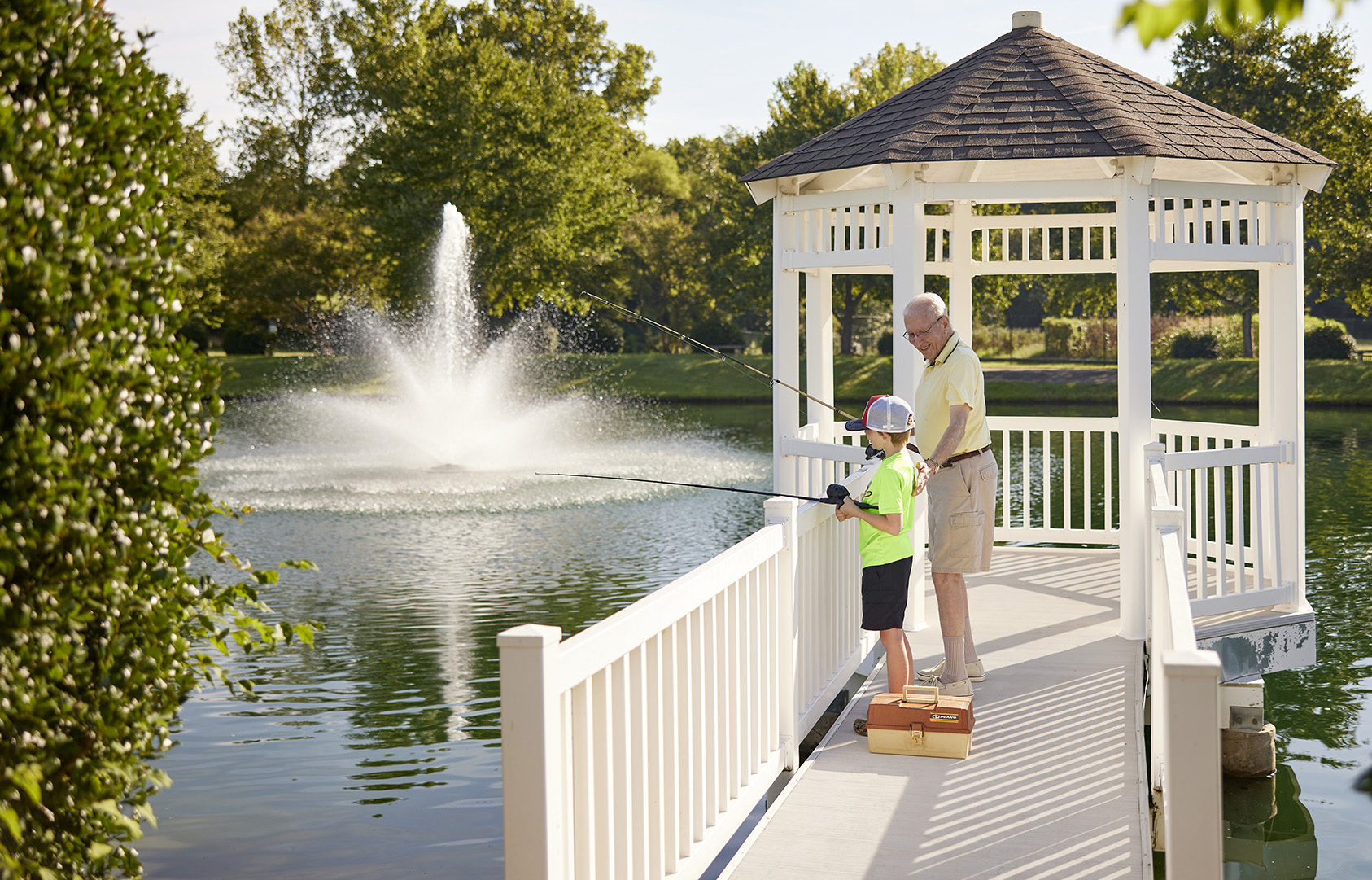 An elderly man and a young boy fishing from a gazebo by a pond with a fountain.
