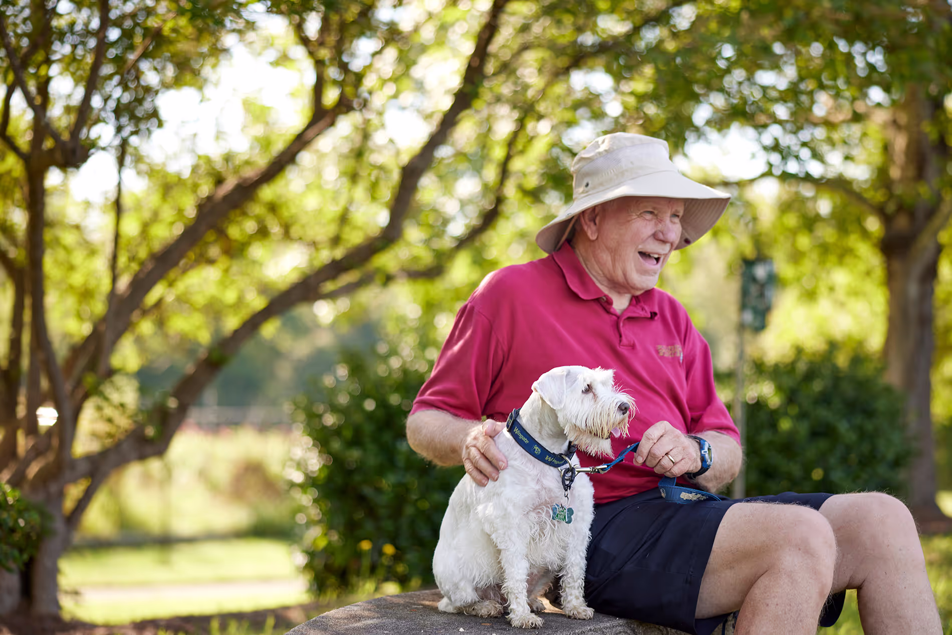 An older man in a hat sits on a bench with a small white dog.