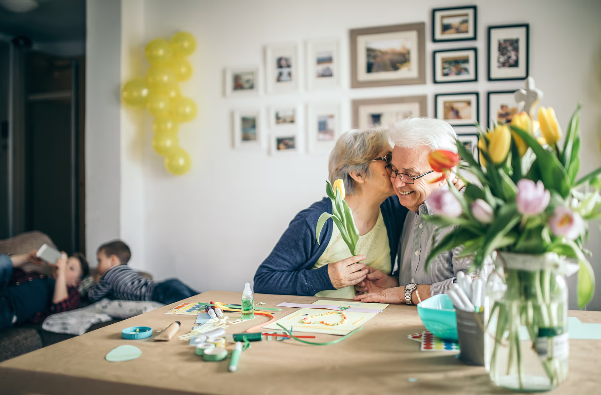 An elderly woman kisses an elderly man on the cheek while holding a tulip.