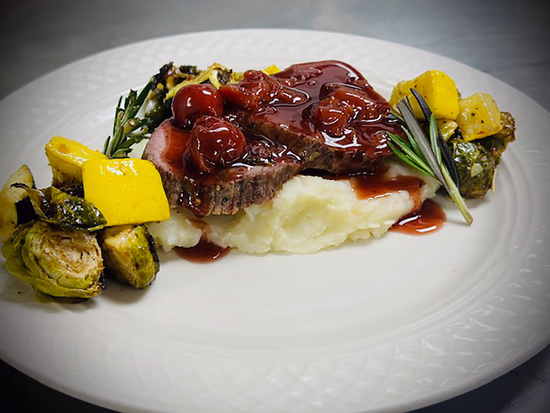 Plate of sliced meat with cherry sauce, mashed potatoes, roasted vegetables, and a rosemary garnish.
