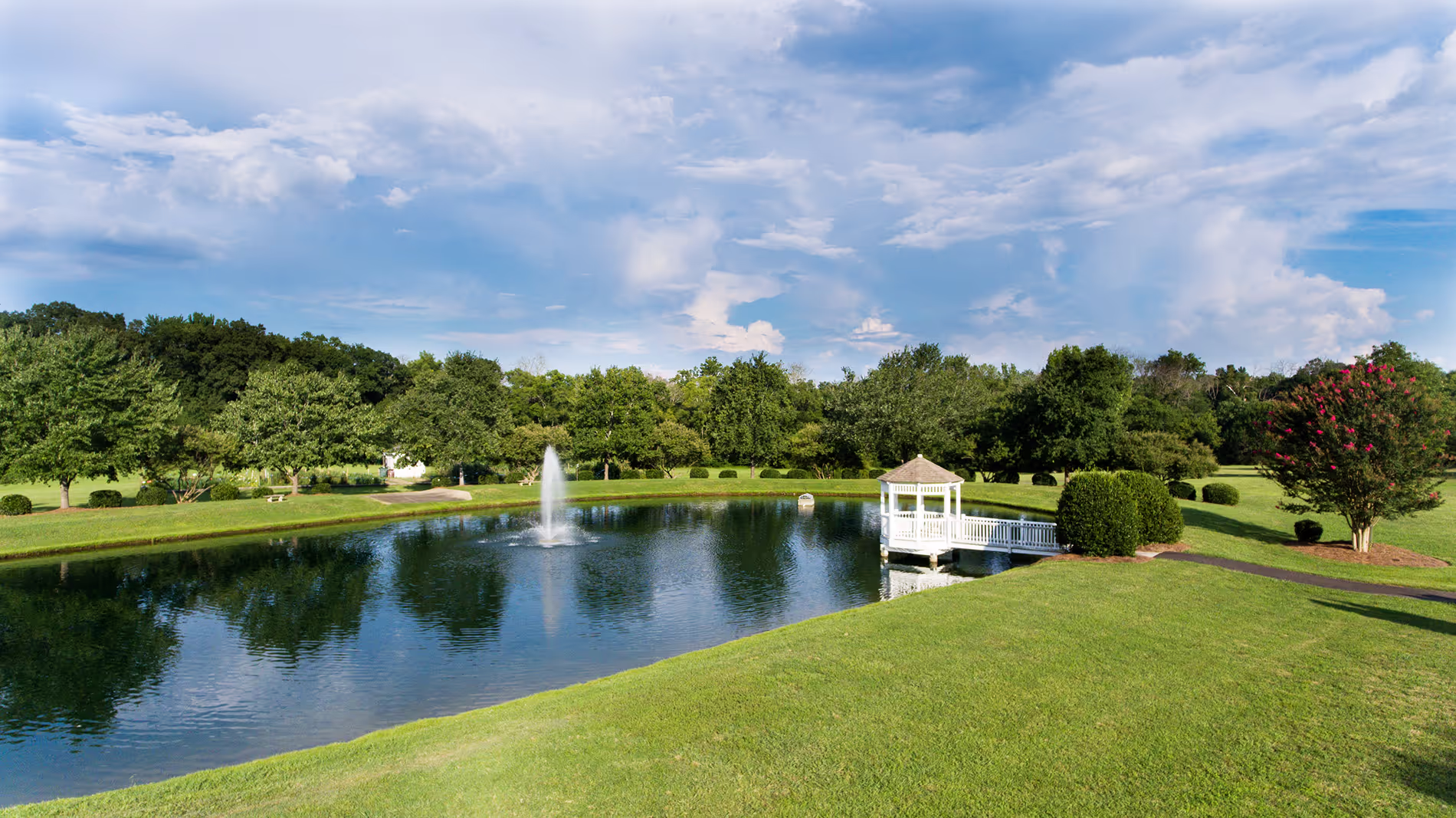 A small pond with a fountain and a small white gazebo surrounded by grass and trees.