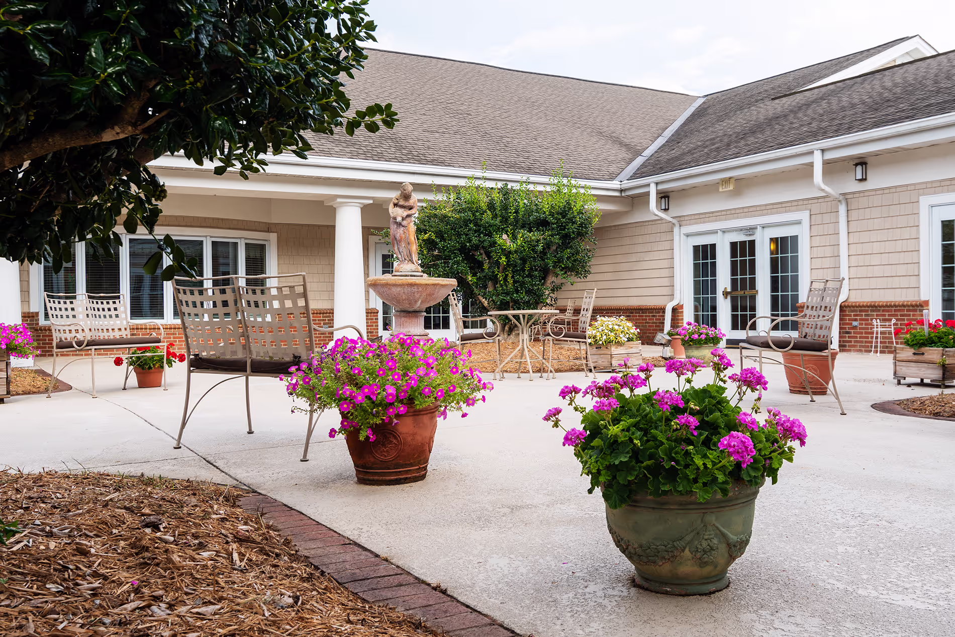 A courtyard with chairs and a statue surrounded by potted plants with pink flowers.