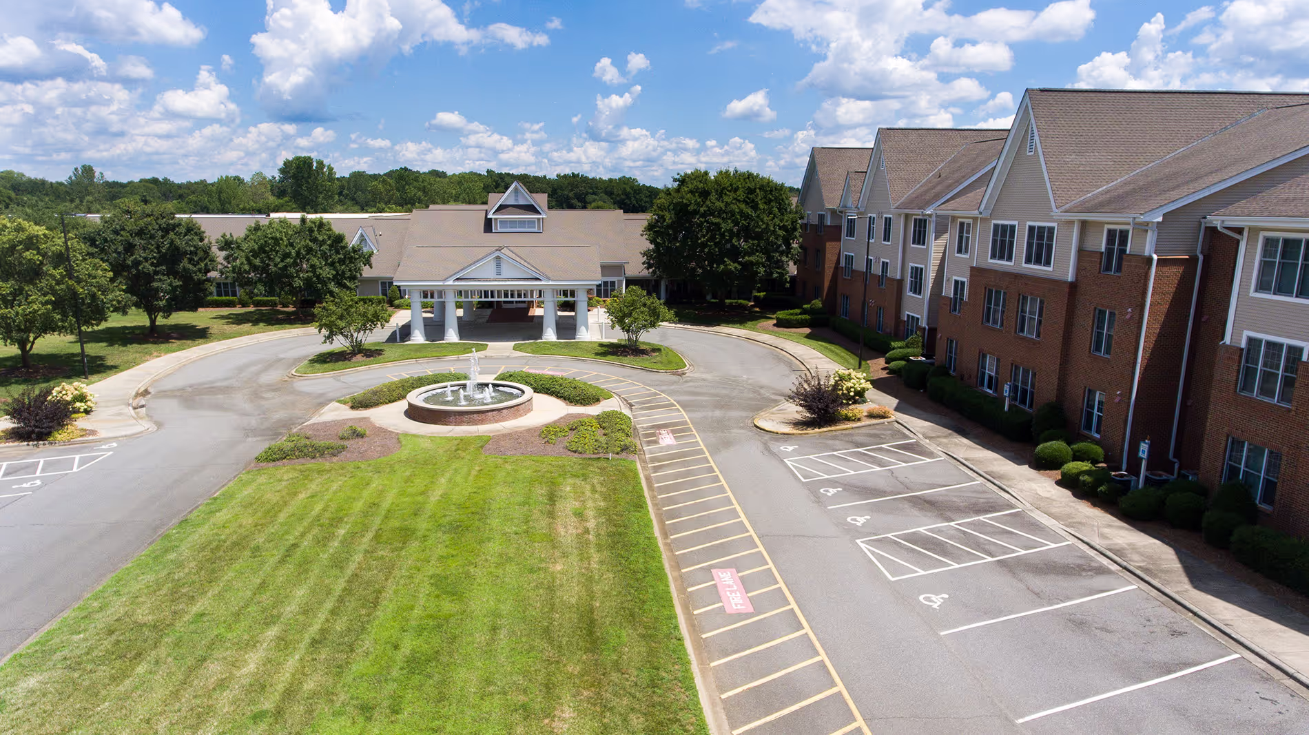 Aerial view of a large building with a circular driveway and fountain, surrounded by greenery and parking spaces.