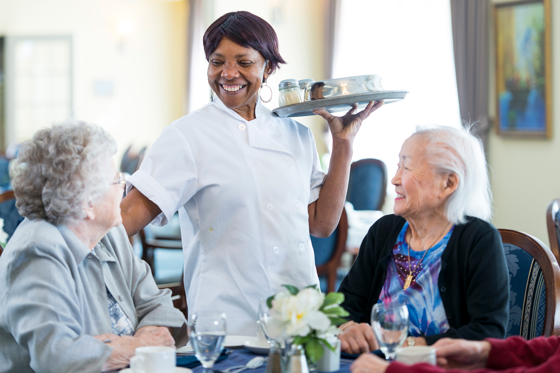 A smiling server carries a tray while assisting elderly women seated at a dining table.