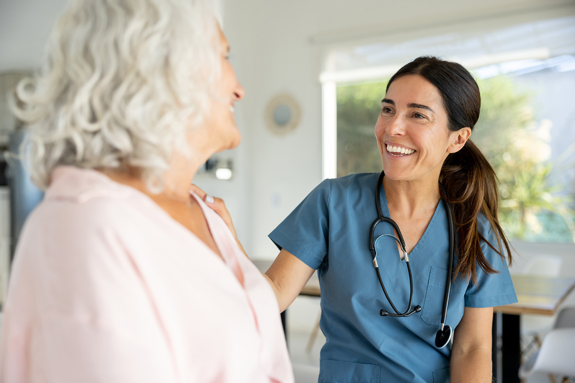 A nurse with a stethoscope talks and smiles with a patient in a room.