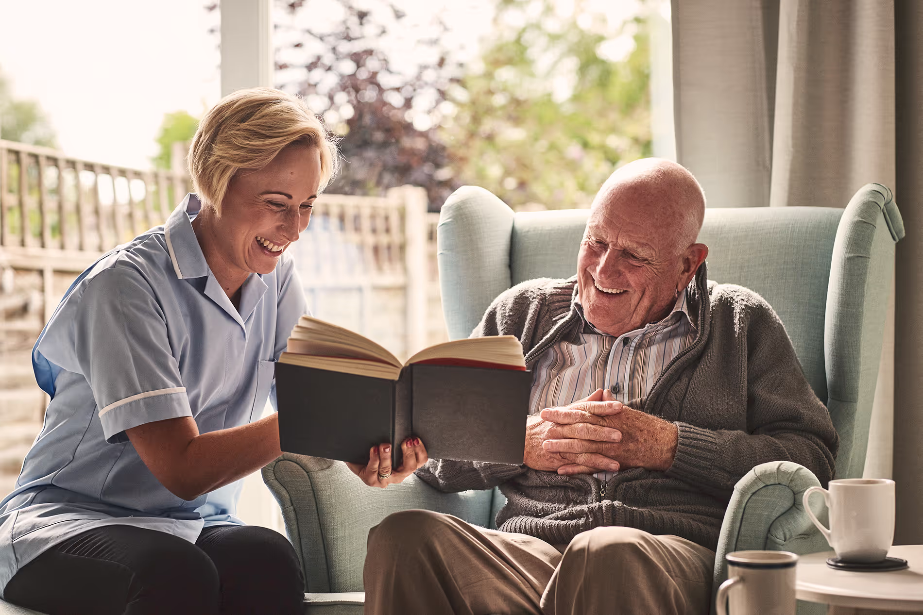 A caregiver and an elderly man sit together, smiling and reading a book in a cozy room.